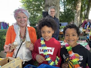 Flohmarkt 2025 Im Vordergrund links eine Frau in oranger Strickjacke, daneben zwei dunkelhäutige Kinder, ein Mädchen und ein Junge. Im Hintergrund ein älterer freundlicher Mann, der in die Kamera lächelt.